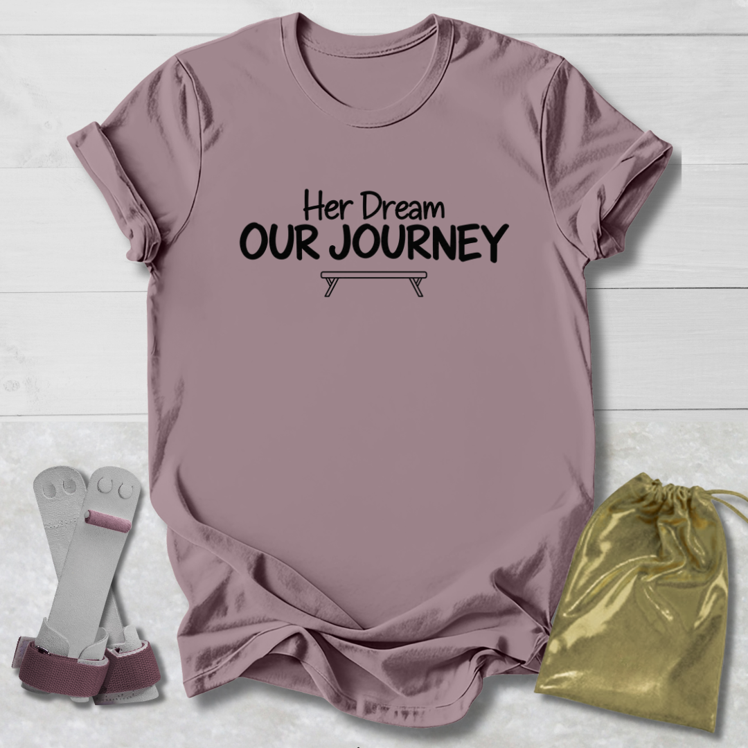 A dusty mauve short-sleeve t-shirt with the black printed text 'Her Dream OUR JOURNEY' and a small bench graphic, laid flat on a white wood background. A pair of gray and mauve athletic support sleeves and a shiny gold drawstring pouch are placed beside it.