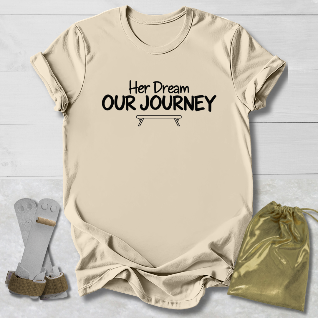 A beige graphic t-shirt with the text 'Her Dream OUR JOURNEY' and a small tent illustration, displayed on a white wood background alongside a pair of gray and brown hiking boots and a gold drawstring pouch.