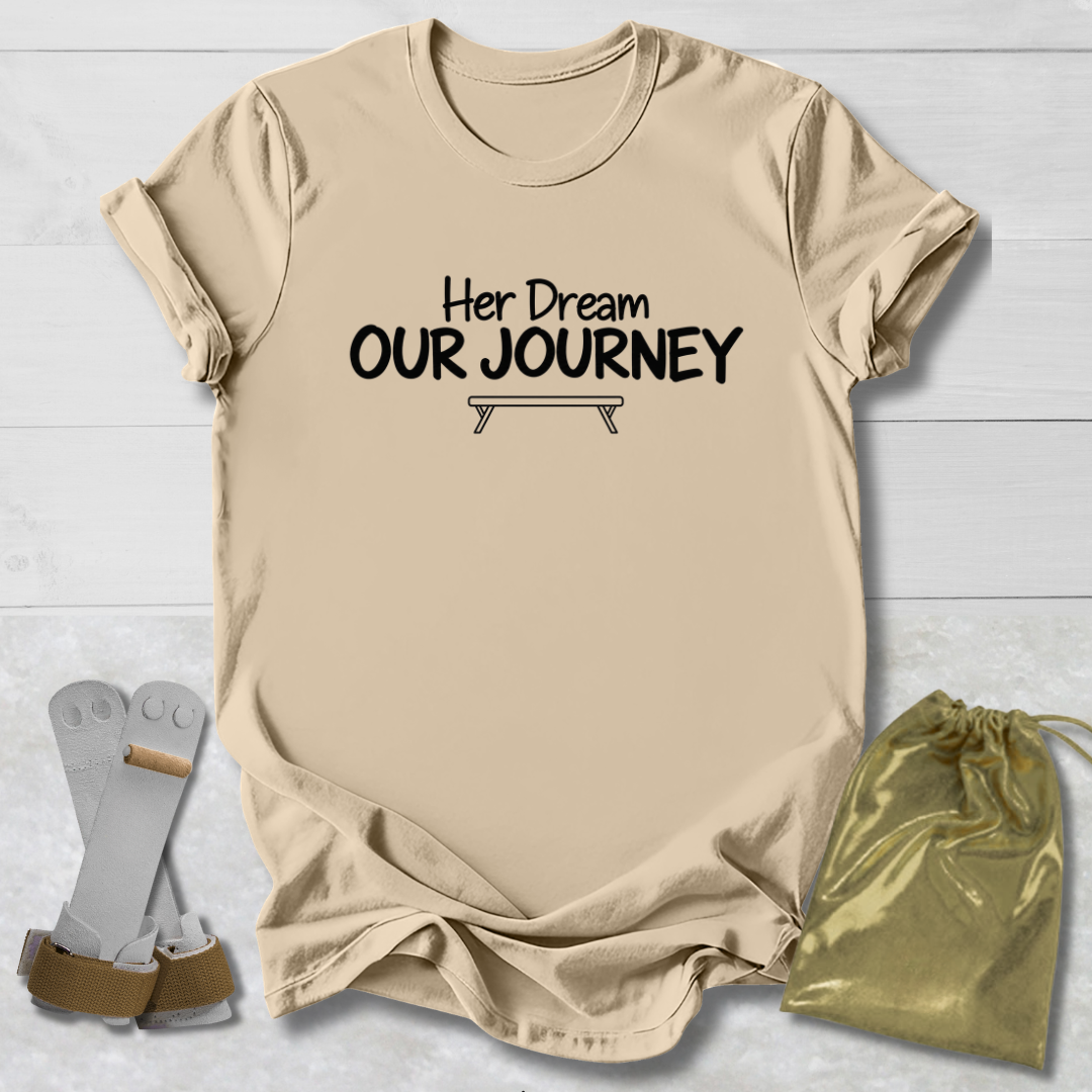 A beige graphic t-shirt with the text 'Her Dream OUR JOURNEY' and a small tent illustration, displayed on a white wood background with a pair of white and brown sandals and a gold drawstring pouch.
