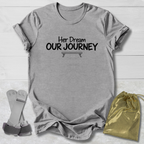 A heather gray short-sleeve t-shirt with the black printed text 'Her Dream OUR JOURNEY' and a small bench graphic, laid flat on a white wood background. A gold drawstring pouch and a pair of gray straps are placed beside it.