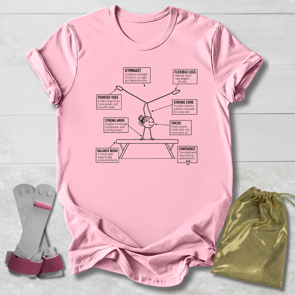 A light pink graphic t-shirt featuring a gymnast diagram with labeled body parts and words like 'Gymnast', 'Flexible Legs', and 'Strong Core'. The shirt is displayed flat on a light gray wood background, accompanied by a pair of pink and gray gymnastic straps and a shiny gold drawstring bag.