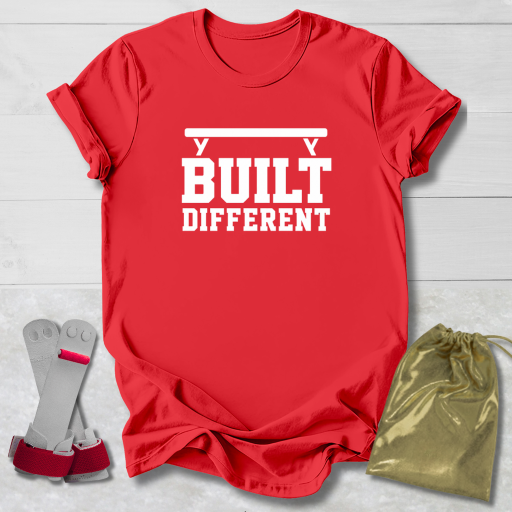 A red short-sleeve T-shirt with bold white text reading 'BUILT DIFFERENT' and a barbell graphic above, shown on a light wood background with a pair of wrist straps and a small gold drawstring pouch as coordinating accessories.