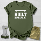An olive green graphic t-shirt with the white text 'BUILT DIFFERENT' and a barbell graphic, displayed on a white wood background alongside a pair of white and green weightlifting straps and a gold drawstring bag.