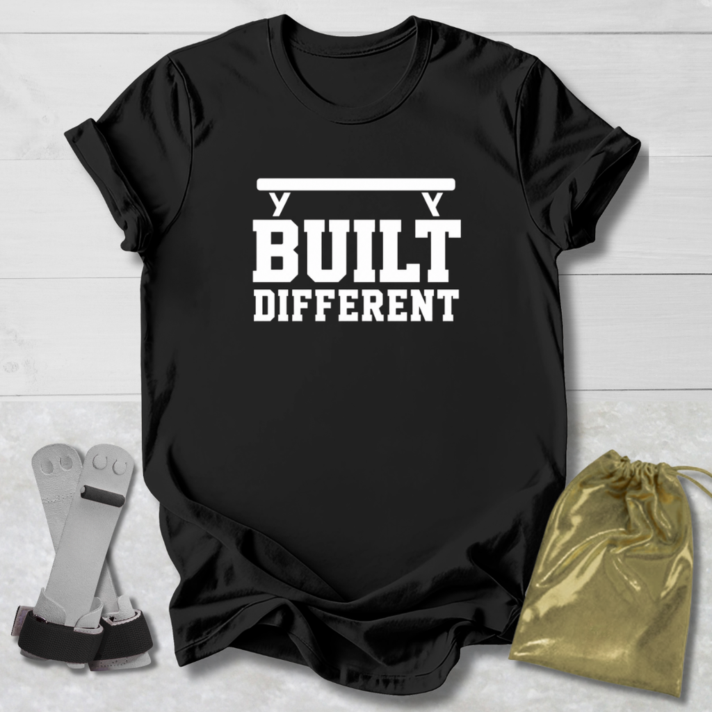 A black short-sleeve graphic T-shirt laid flat on a light wood background, with white text reading 'BUILT DIFFERENT' and a barbell graphic above. A pair of gray and black wrist wraps and a small metallic gold drawstring pouch are placed beside the shirt as styling props.