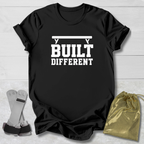 A black short-sleeve graphic T-shirt laid flat on a light wood background, with white text reading 'BUILT DIFFERENT' and a barbell graphic above. A pair of gray and black wrist wraps and a small metallic gold drawstring pouch are placed beside the shirt as styling props.