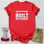 A red short-sleeve T-shirt with bold white text reading 'BUILT DIFFERENT' and a barbell graphic above, shown on a light wood background with a pair of wrist straps and a small gold drawstring pouch as coordinating accessories.