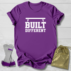 A purple short-sleeve graphic T-shirt laid flat on a light wood background, with white text reading 'BUILT DIFFERENT' and a barbell graphic above. A pair of purple wrist wraps and a gold drawstring pouch are placed beside the shirt as included accessories.