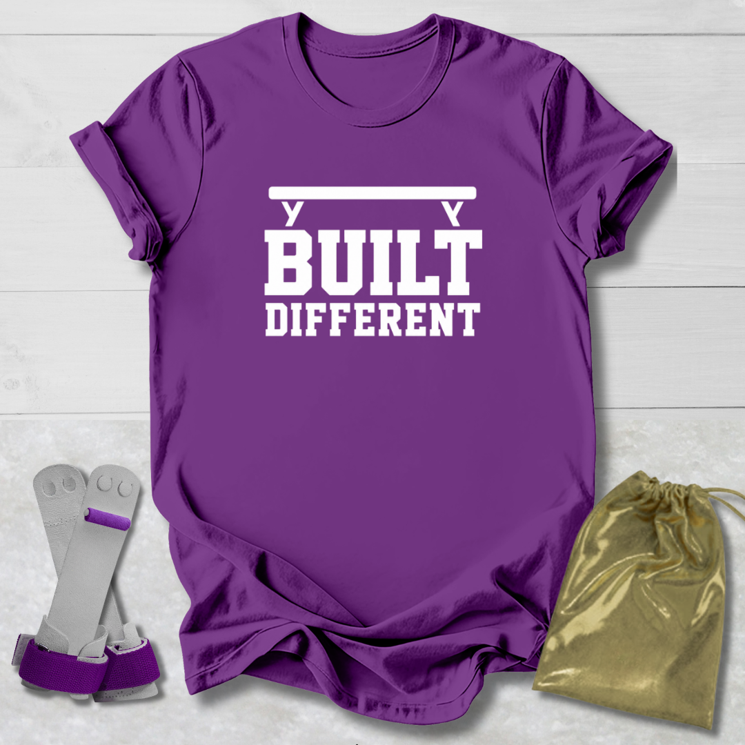 A purple short-sleeve graphic T-shirt laid flat on a light wood background, with white text reading 'BUILT DIFFERENT' and a barbell graphic above. A pair of purple wrist wraps and a gold drawstring pouch are placed beside the shirt as included accessories.