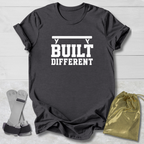 A dark charcoal gray t-shirt with a white graphic reading 'BUILT DIFFERENT' under a barbell, styled on a white wood background with a pair of weight plates and a gold drawstring bag.