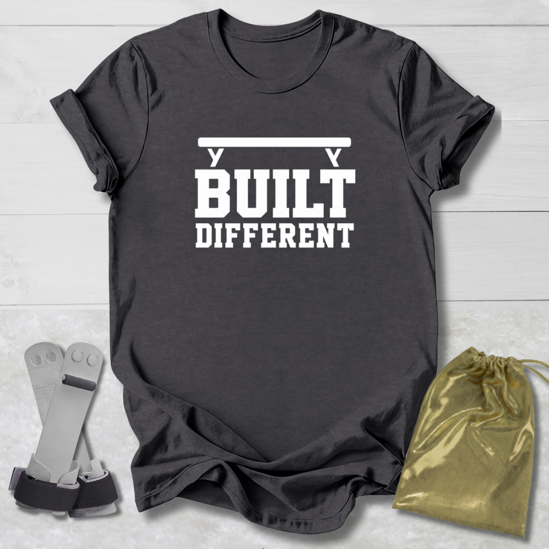 A dark charcoal gray t-shirt with a white graphic reading 'BUILT DIFFERENT' under a barbell, styled on a white wood background with a pair of weight plates and a gold drawstring bag.