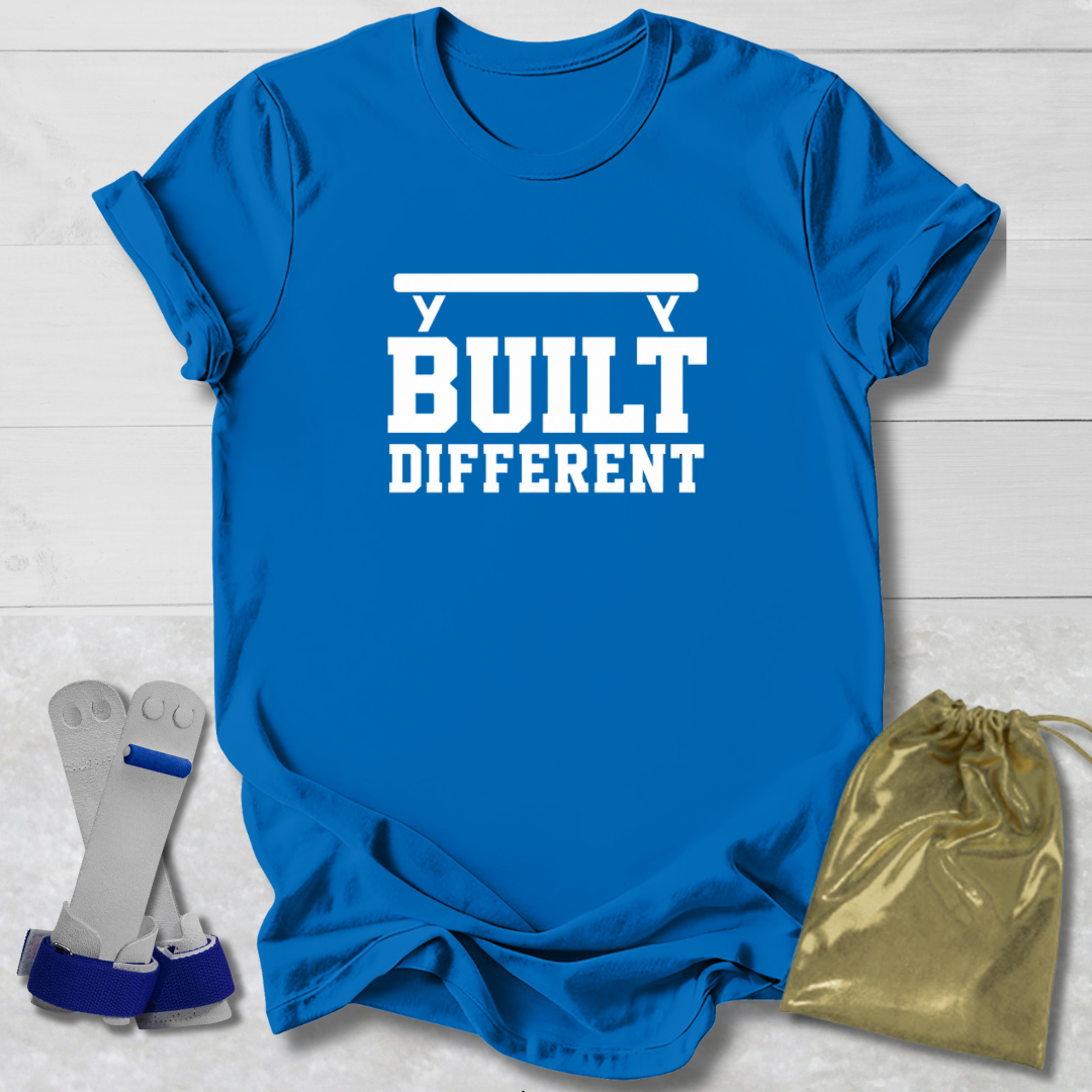 A bright blue short-sleeve T-shirt laid flat on a white wood background, printed with the phrase 'BUILT DIFFERENT' in white block letters with a bar graphic above. A pair of gray and blue wrist wraps and a small metallic gold drawstring pouch are placed beside the shirt as styling props.