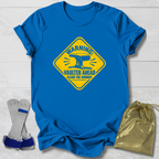 A royal blue short-sleeve T-shirt laid flat on a white wood background, featuring a large yellow warning-style graphic with an anvil and the words 'WARNING Vaulter Ahead Clear the Runway.' Two gray-and-blue weight plates and a shiny gold drawstring pouch are placed beside the shirt as styling props.