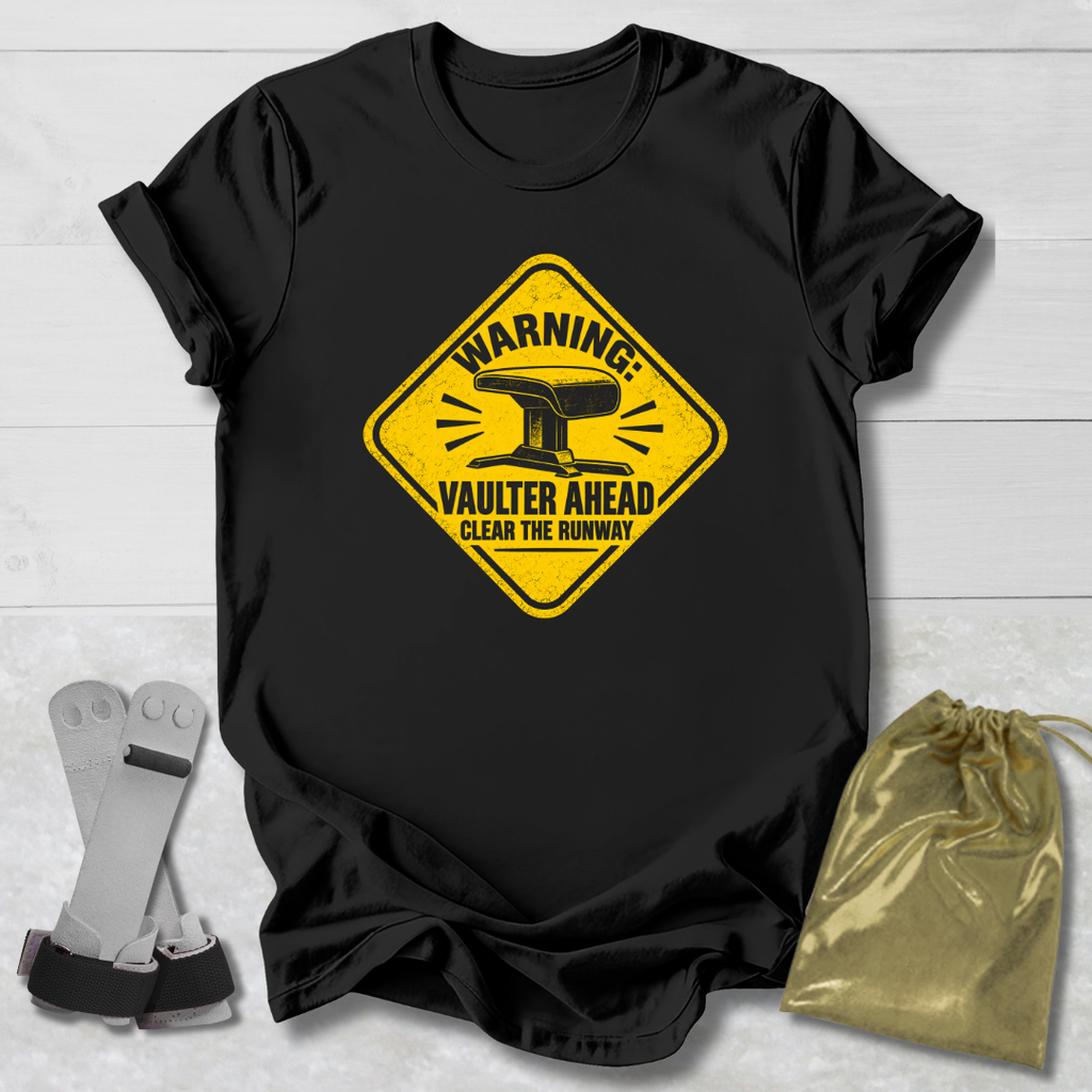 A black short-sleeve graphic T-shirt laid flat on a light wood background, featuring a yellow warning-sign style print with an anvil and the text 'WARNING VAULTER AHEAD CLEAR THE RUNWAY.' A metallic gold drawstring pouch and a pair of gray-and-black gloves are placed beside the shirt as styling props.