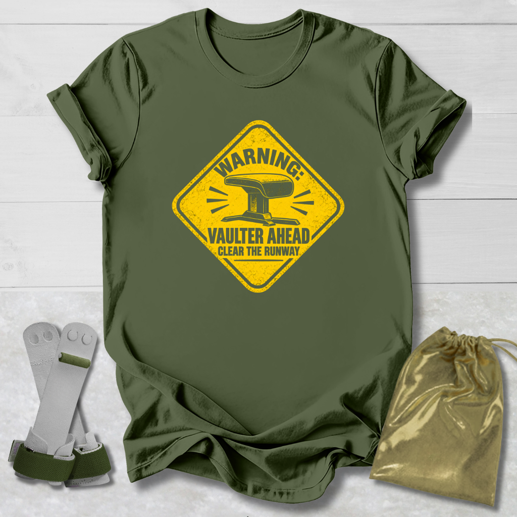 An olive green short-sleeve t-shirt with a yellow warning sign graphic that reads 'WARNING: VAULTER AHEAD CLEAR THE RUNWAY' with an anvil illustration. The shirt is displayed flat on a white wood background alongside a pair of white and green metal tool brackets and a shiny gold drawstring pouch.