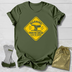An olive green short-sleeve t-shirt with a yellow warning sign graphic that reads 'WARNING: VAULTER AHEAD CLEAR THE RUNWAY' with an anvil illustration. The shirt is displayed flat on a white wood background alongside a pair of white and green metal tool brackets and a shiny gold drawstring pouch.