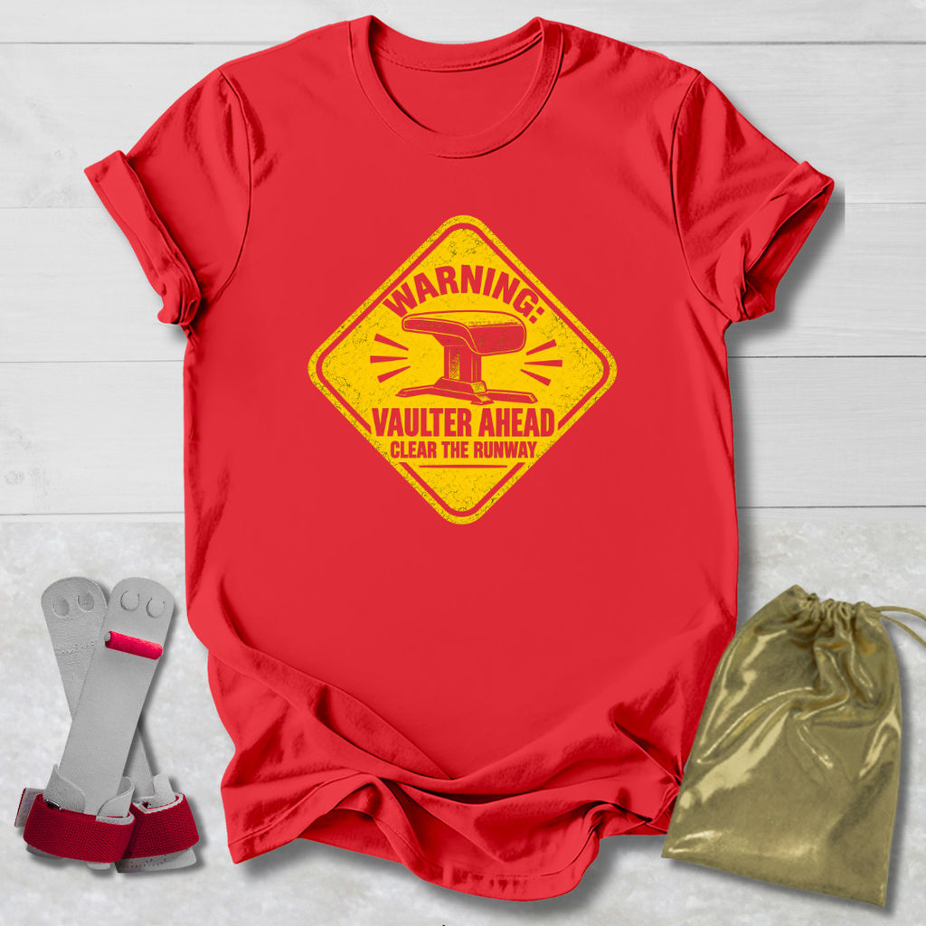 A red short-sleeve crewneck t-shirt with a yellow warning sign graphic that reads 'WARNING! VAULTER AHEAD CLEAN THE RUNWAY'. The shirt is displayed on a white wood background alongside a pair of gray and red wing-like accessories and a gold drawstring pouch.