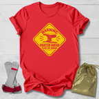 A red short-sleeve crewneck t-shirt with a yellow warning sign graphic that reads 'WARNING! VAULTER AHEAD CLEAN THE RUNWAY'. The shirt is displayed on a white wood background alongside a pair of gray and red wing-like accessories and a gold drawstring pouch.