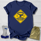 A navy blue graphic t-shirt laid flat on a white wood background, featuring a yellow warning sign design with an anvil and the text 'WARNING: VAULTER AHEAD CLEAR THE RUNWAY'. To the left of the shirt are white and blue roller skate blades, and to the right is a shiny gold drawstring bag.