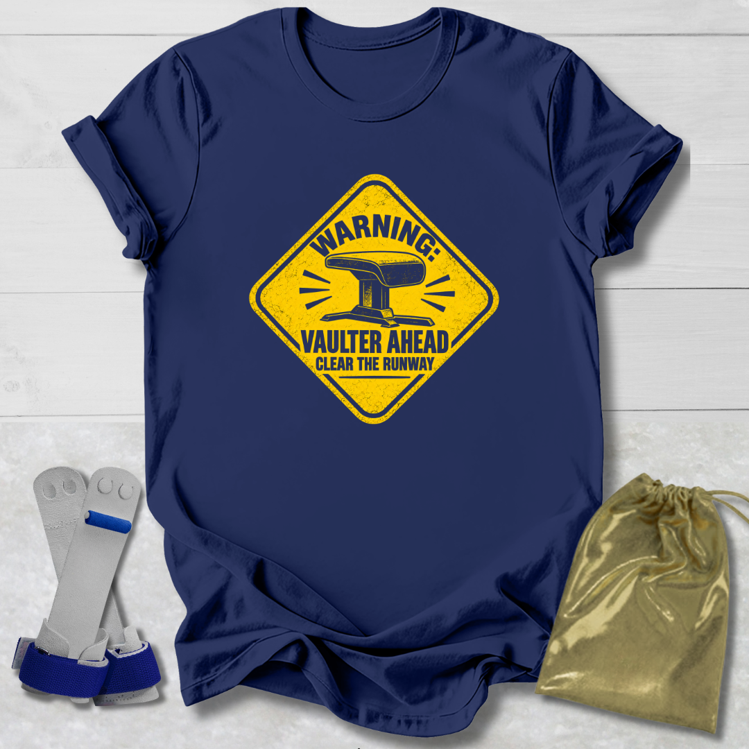 A navy blue graphic t-shirt laid flat on a white wood background, featuring a yellow warning sign design with an anvil and the text 'WARNING: VAULTER AHEAD CLEAR THE RUNWAY'. To the left of the shirt are white and blue roller skate blades, and to the right is a shiny gold drawstring bag.