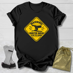A black short-sleeve graphic T-shirt laid flat on a light wood background, featuring a yellow warning-sign style print with an anvil and the text 'WARNING VAULTER AHEAD CLEAR THE RUNWAY.' A metallic gold drawstring pouch and a pair of gray-and-black gloves are placed beside the shirt as styling props.