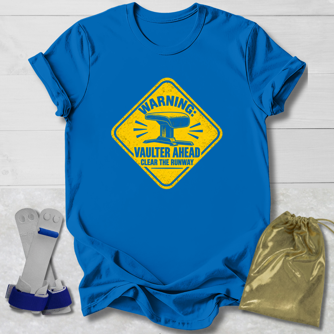 A royal blue short-sleeve T-shirt laid flat on a white wood background, featuring a large yellow warning-style graphic with an anvil and the words 'WARNING Vaulter Ahead Clear the Runway.' Two gray-and-blue weight plates and a shiny gold drawstring pouch are placed beside the shirt as styling props.