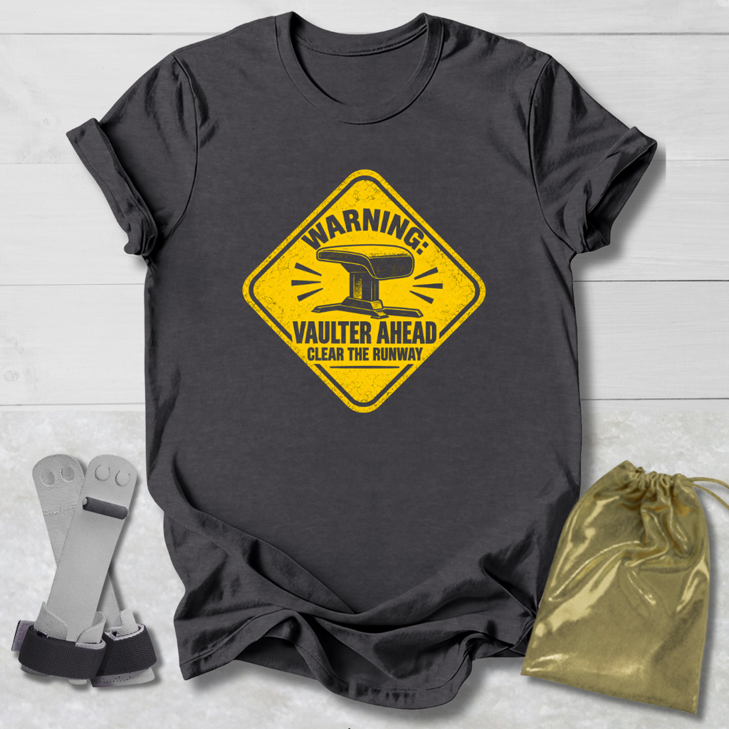 A dark charcoal gray t-shirt with a yellow warning sign graphic that reads 'WARNING: VAULTER AHEAD CLEAR THE RUNWAY' with an anvil illustration. The shirt is displayed flat on a white wood background alongside a pair of gray and black claw tools and a shiny gold drawstring bag.