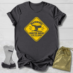 A dark charcoal gray t-shirt with a yellow warning sign graphic that reads 'WARNING: VAULTER AHEAD CLEAR THE RUNWAY' with an anvil illustration. The shirt is displayed flat on a white wood background alongside a pair of gray and black claw tools and a shiny gold drawstring bag.
